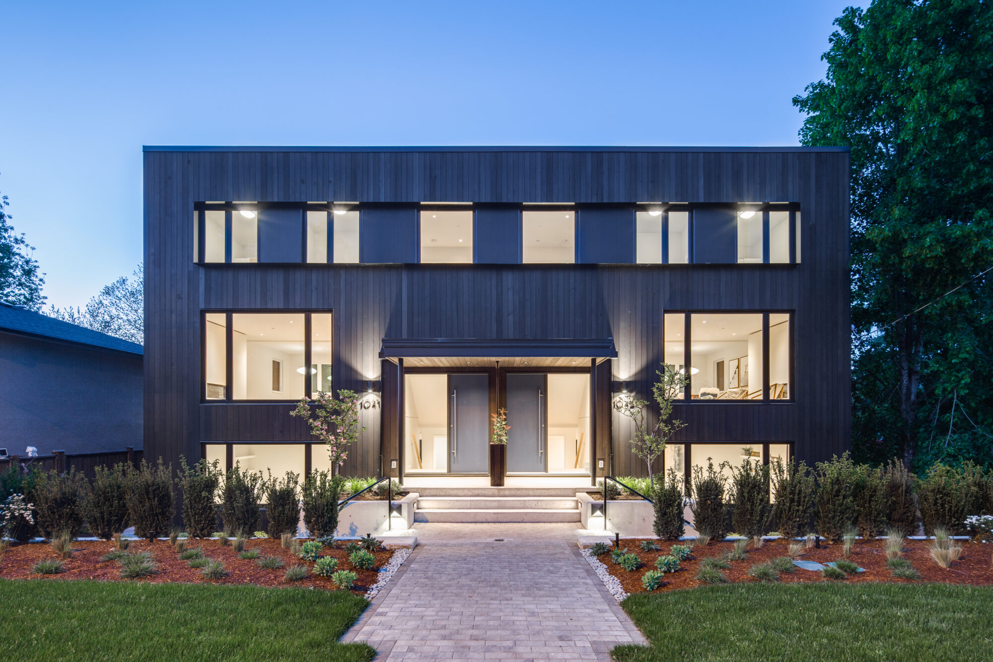 Twilight exterior of modern side-by-side duplex with dark vertical wood siding, blue entry doors and landscaped front yard and entry